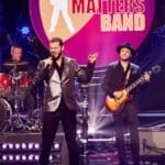 Zanger van de Elvis Matters Band in zilveren colbert zingt op het podium, met gitarist en drummer